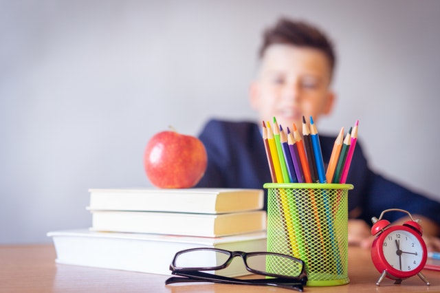 school desk with an apple, books, glasses, and pencils and a young boy out of focus in the background